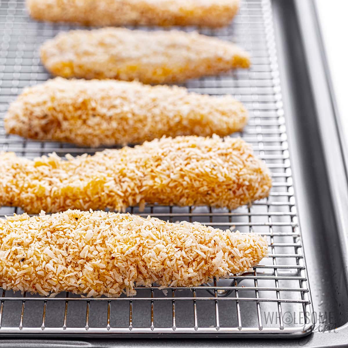 Coated coconut chicken on a rack over a sheet pan, before baking.