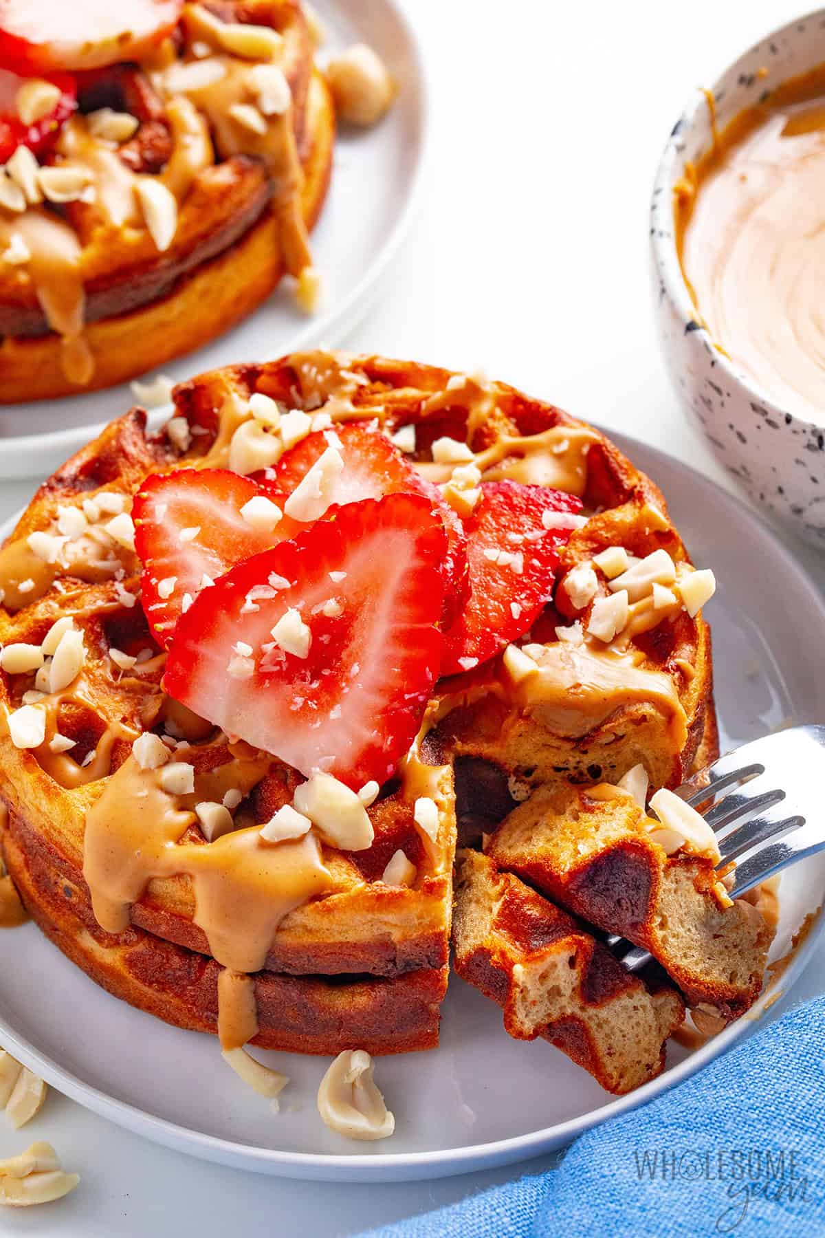 Two plates of golden, fluffy peanut butter waffles topped with fresh fruit and pb drizzle, with a fork lifting a piece.