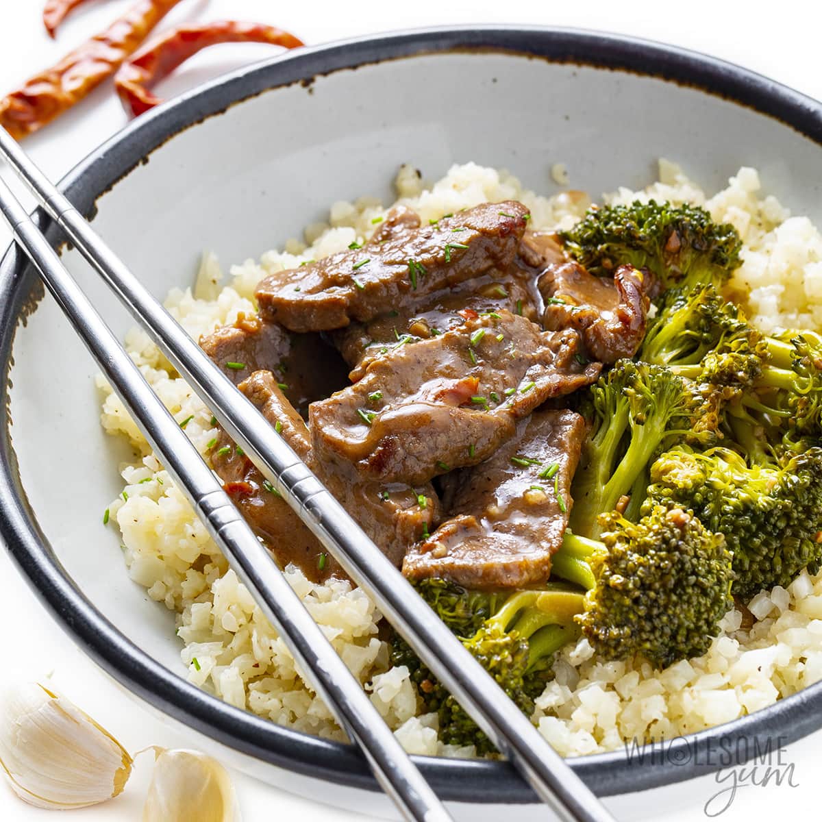 Hunan beef in a bowl with broccoli, cauliflower rice, and chopsticks.
