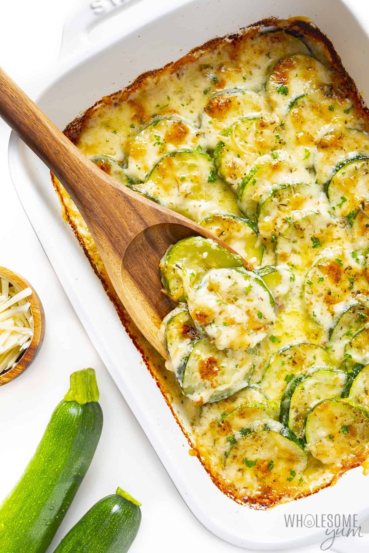 Zucchini gratin with wooden spoon scooping out a portion and fresh zucchini on the side.