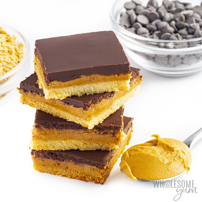 Stack of four keto peanut butter bars with a spoonful of peanut butter and sugar-free chocolate chips in the background