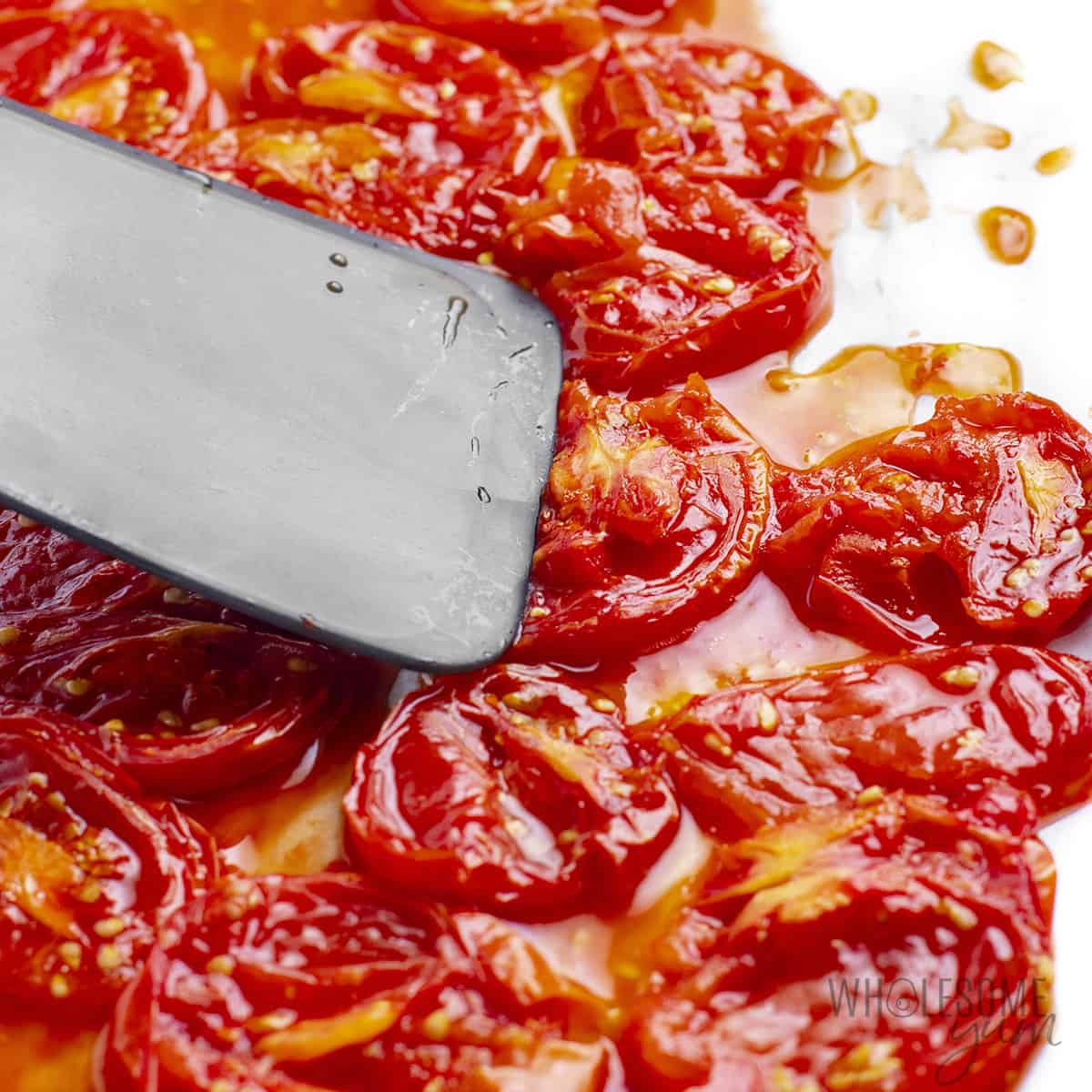 Spatula pressing down on tomatoes in baking sheet.