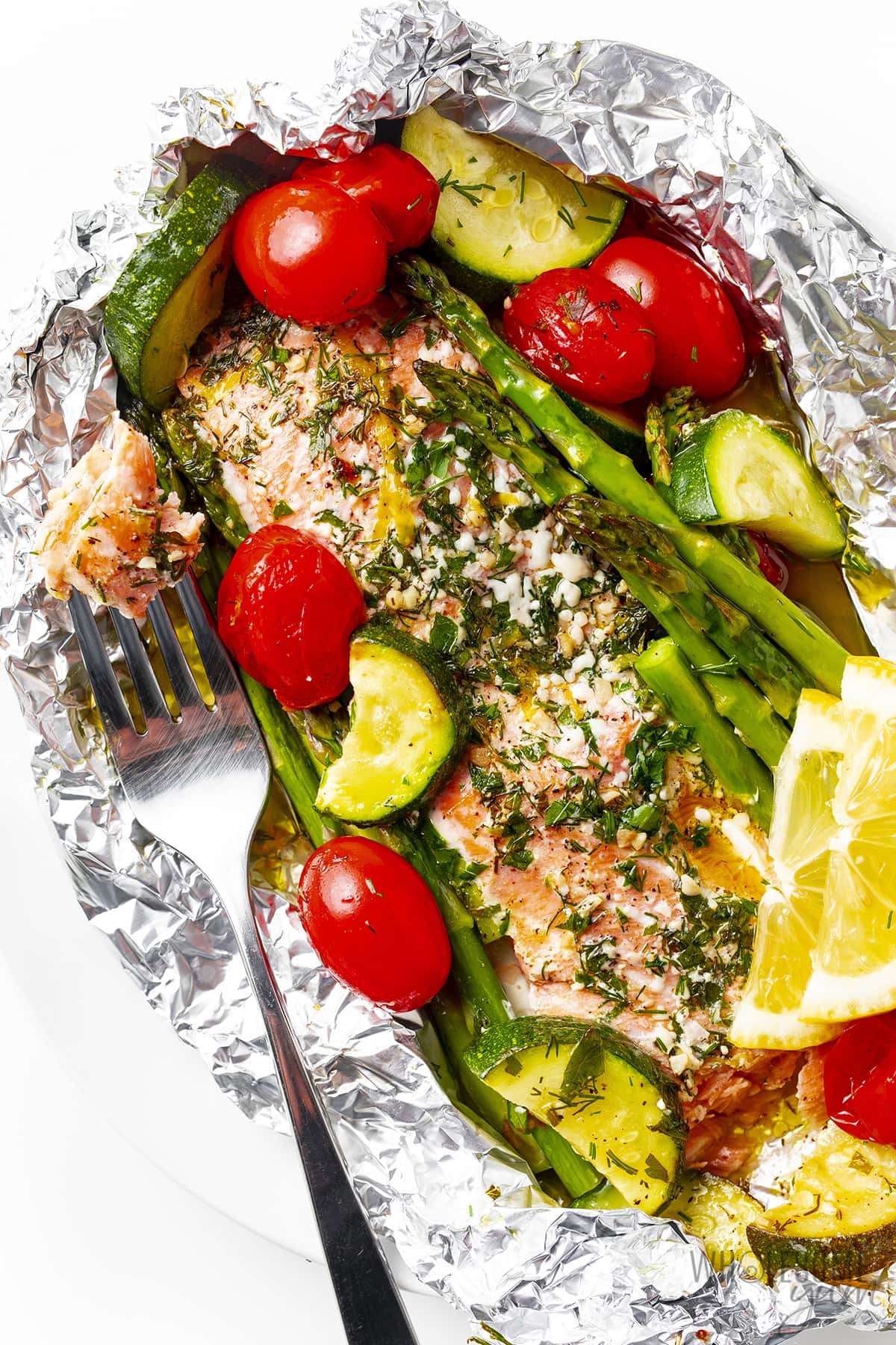 Oven baked salmon in foil with zucchini, asparagus, and tomatoes, and one bite of salmon on a fork.
