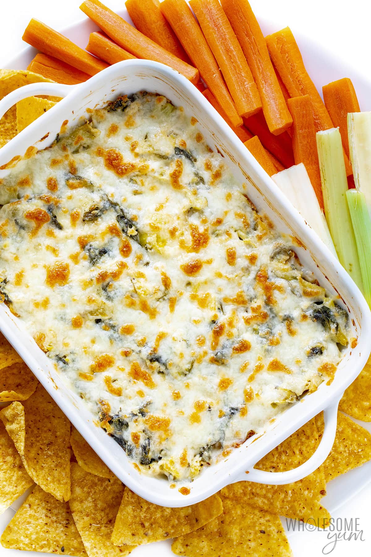 Baked spinach artichoke dip surrounded by carrots, celery, and tortilla chips.