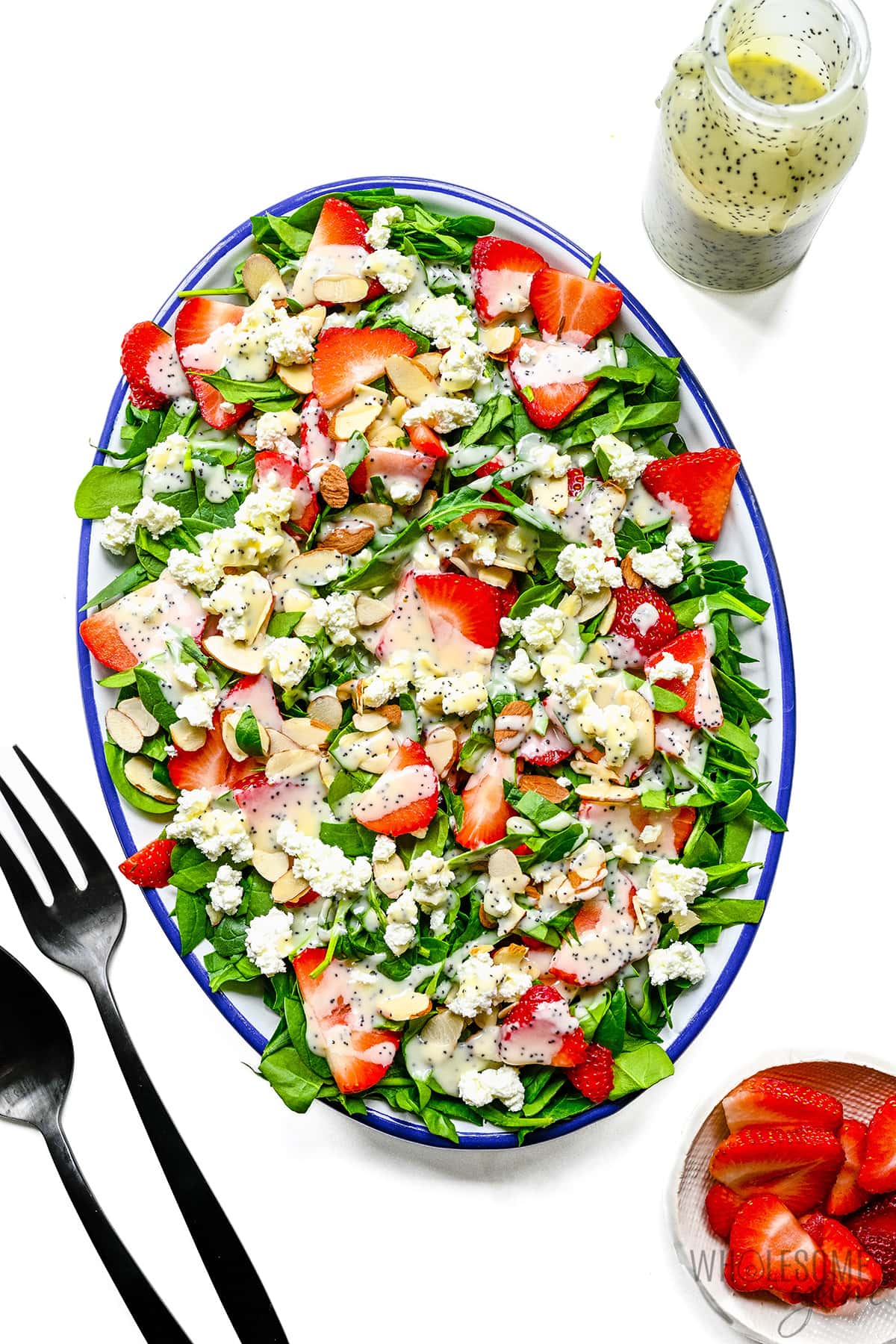 Strawberry spinach salad in a serving bowl next to forks, dressing, and strawberries.