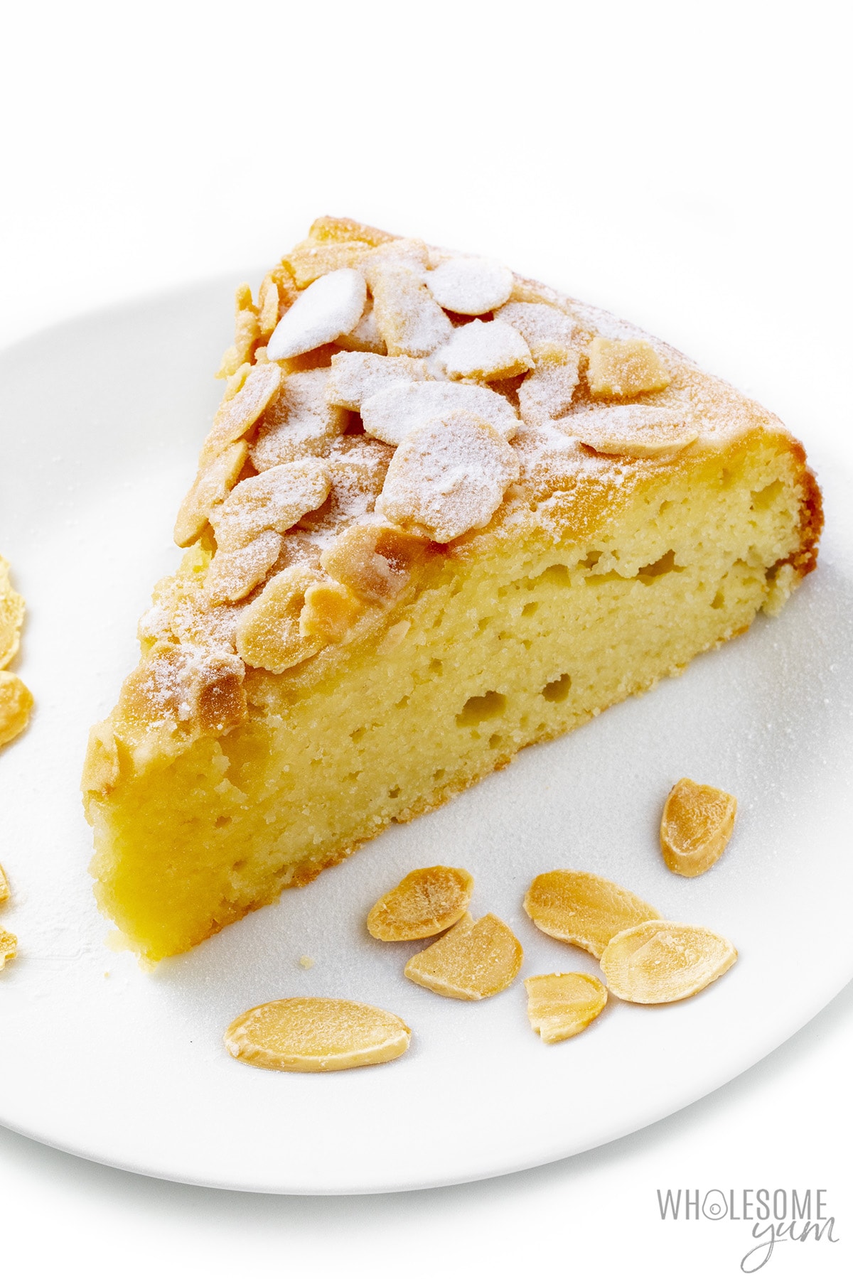 Slice of almond cake on a plate with sliced almonds and a dusting of powdered sweetener.