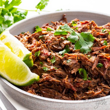 Beef barbacoa recipe on a plate with lime wedges and cilantro.