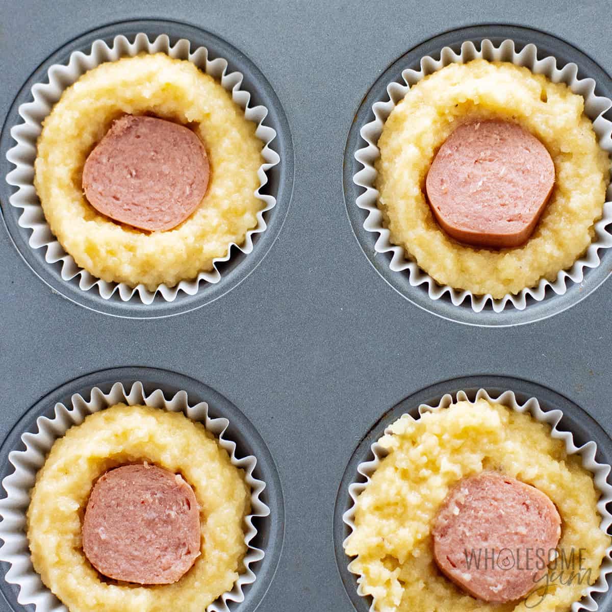 Mini muffin pan filled with cornbread batter, each topped with a piece of hot dog in the center.