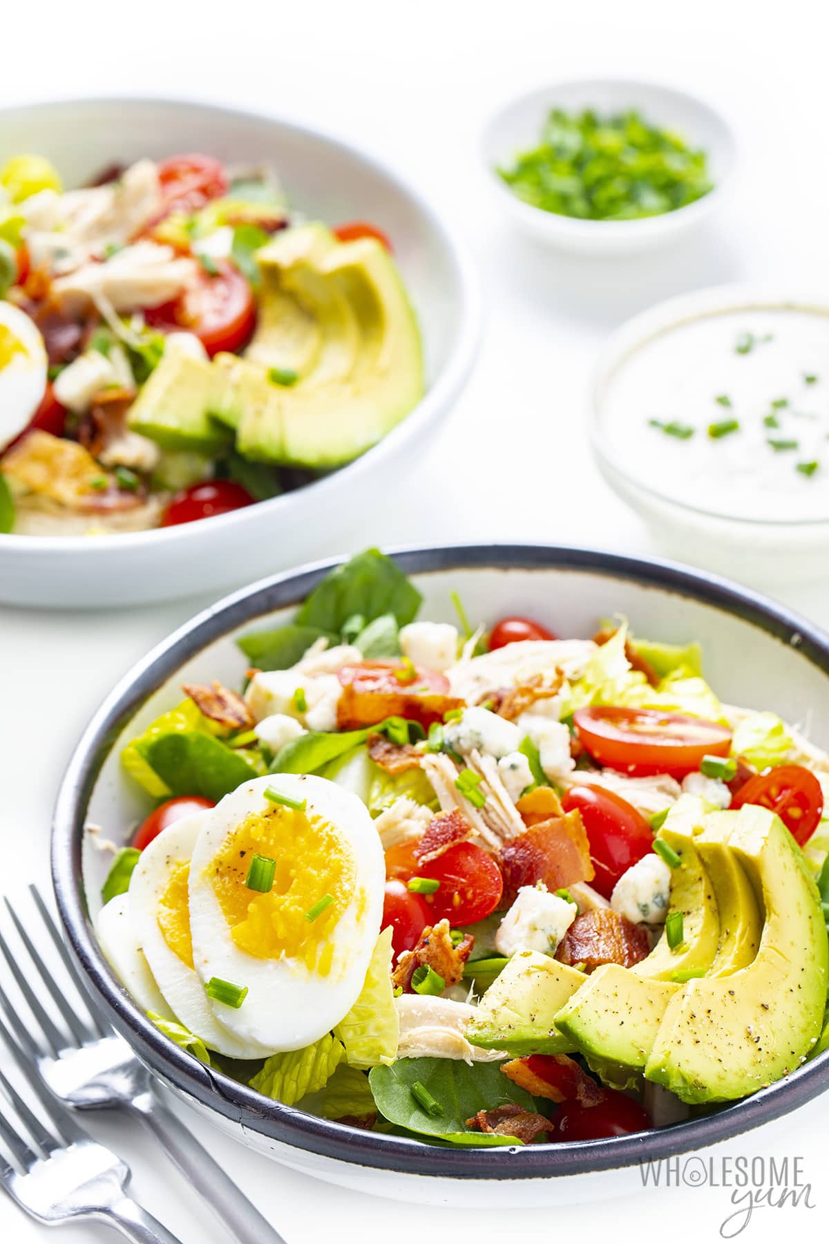 Two bowls of Cobb salad with fork, dressing, and herbs.