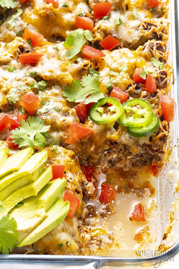 Overhead view of Mexican ground beef casserole in a casserole dish divided into 9 servings with cilantro, jalapenos, and avocado slices and a slice removed
