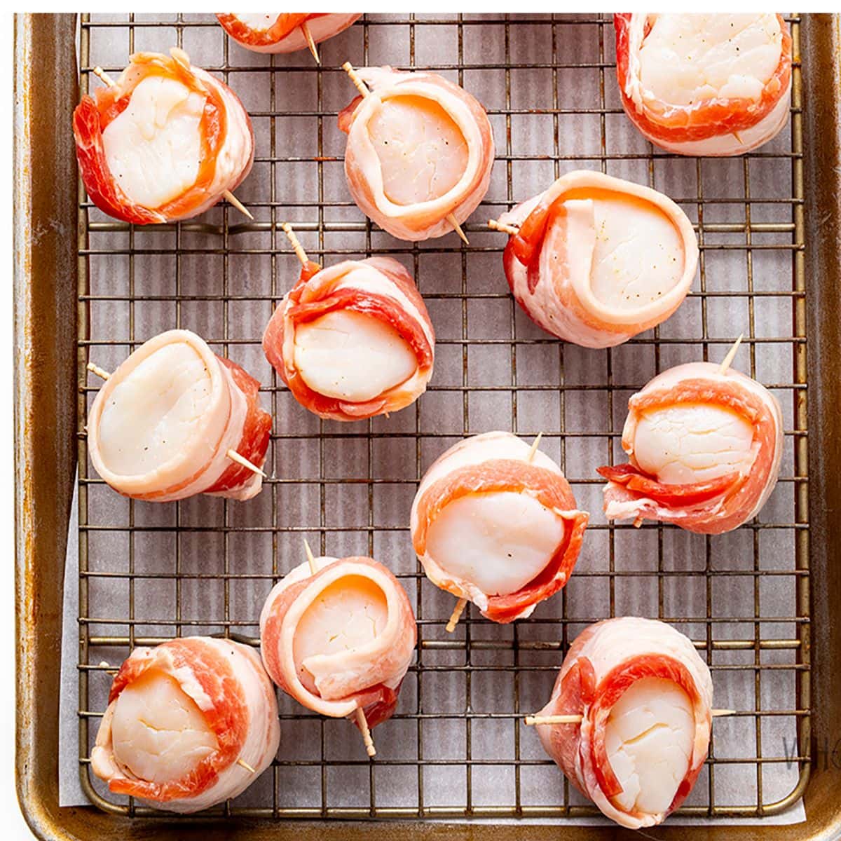 Scallops wrapped in bacon secured with toothpicks and placed on a rack over a sheet pan.