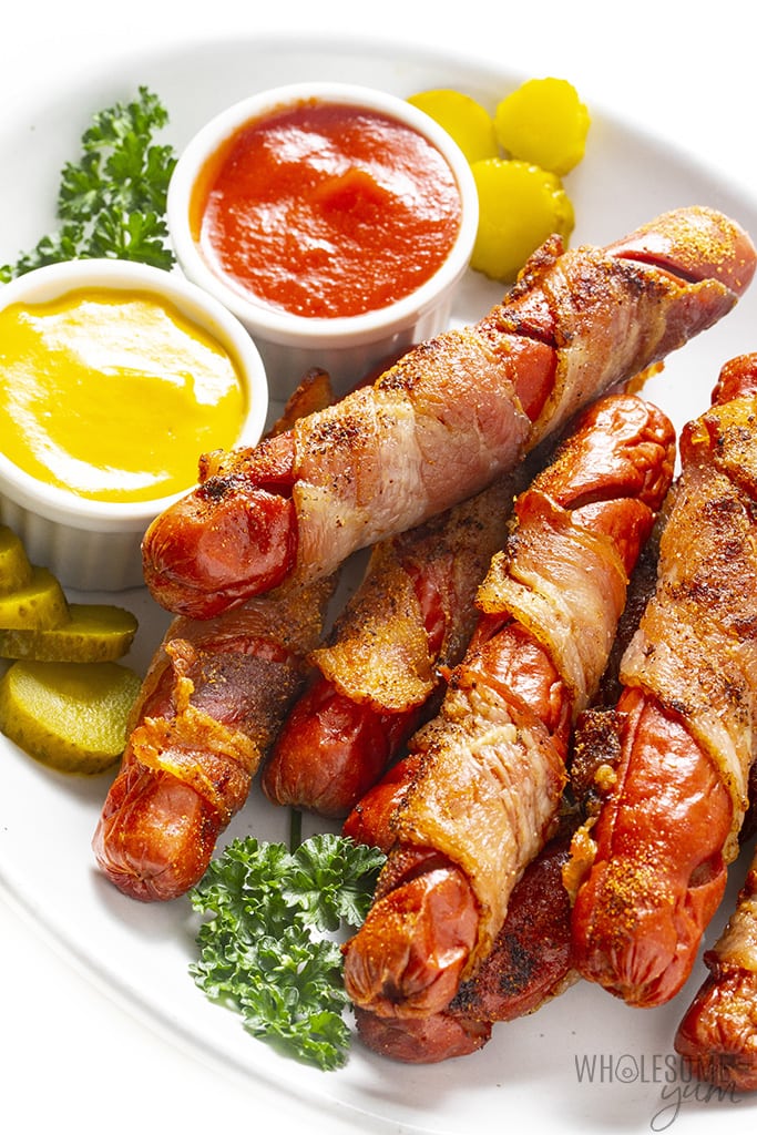 Bacon wrapped hot dogs stacked on a plate with condiments