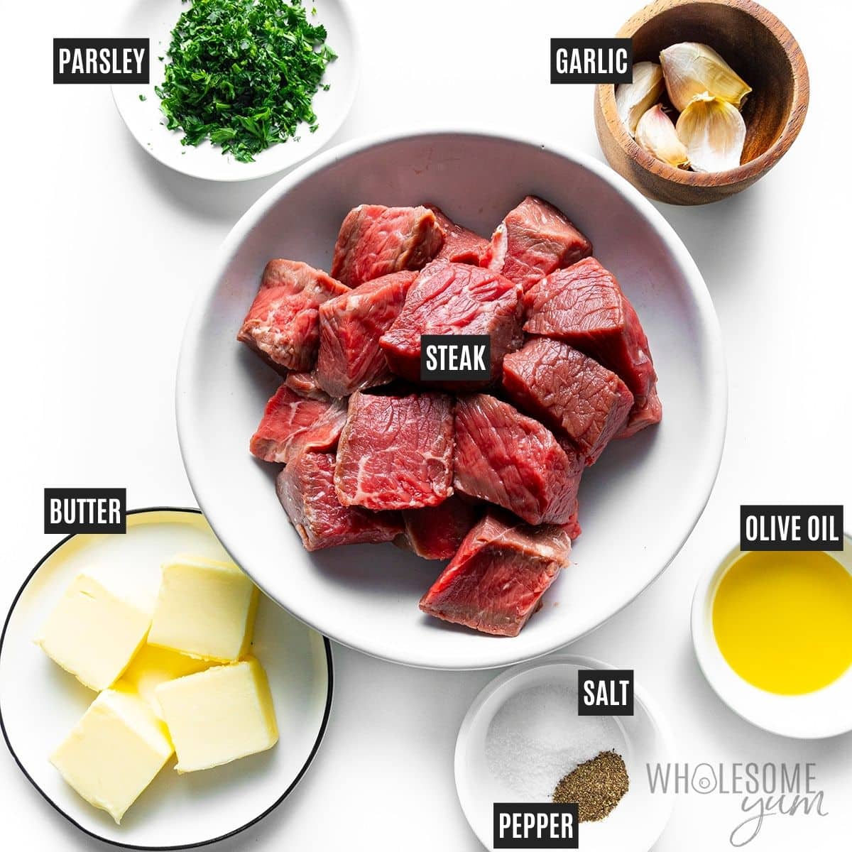 Steak bites in a bowl next to small labeled bowls of butter, garlic, parsley, olive oil, salt, and pepper.