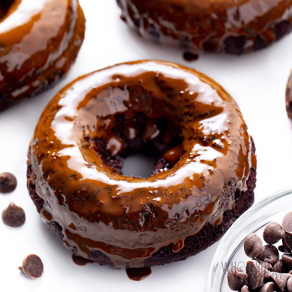 Glazed chocolate protein donuts on a white counter next to some chocolate chips.