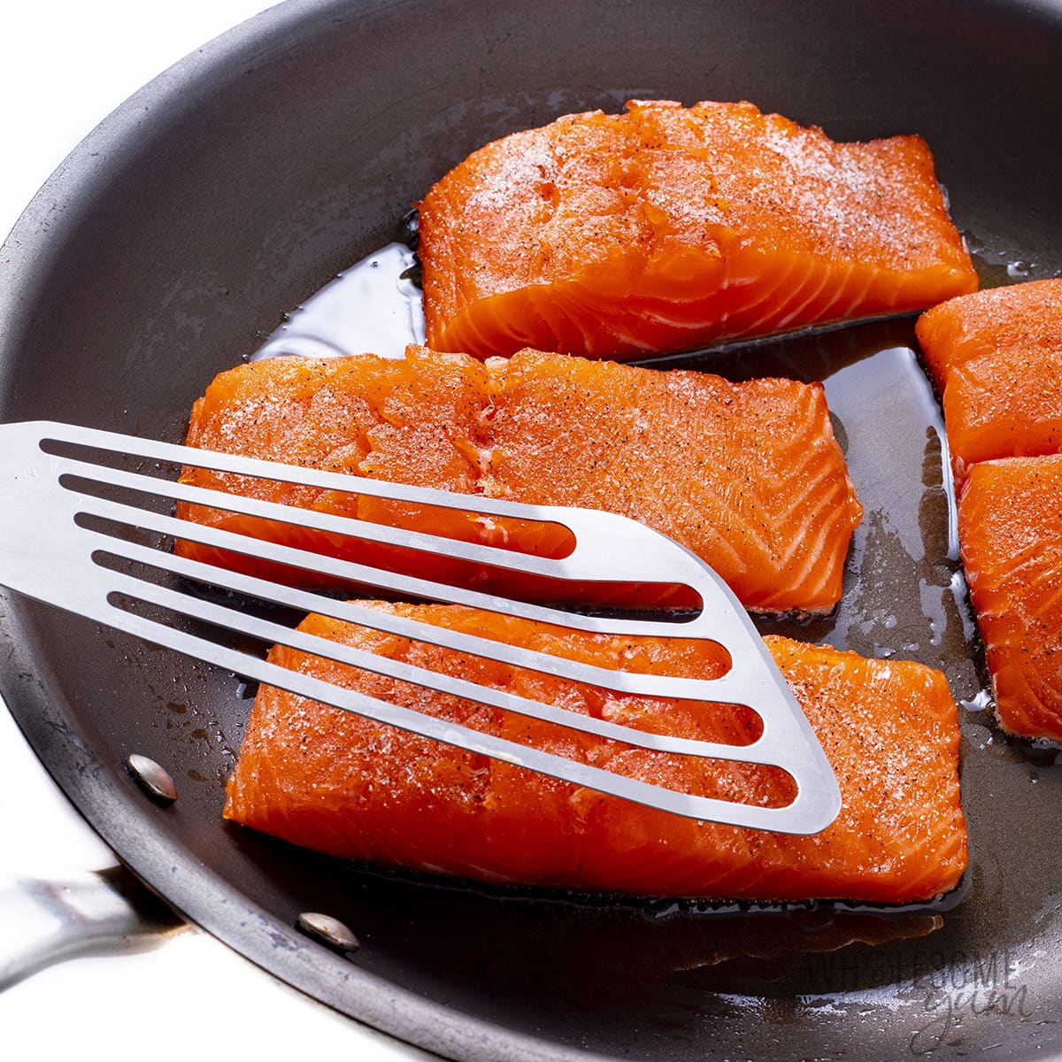 Salmon fillets searing in pan with fish spatula pressing one fillet down.