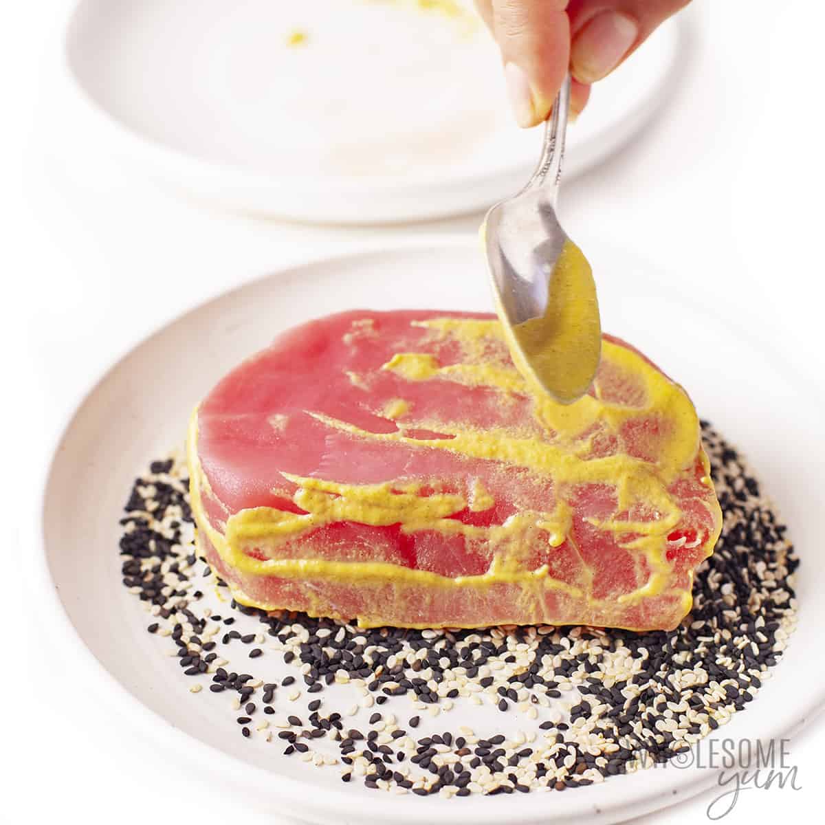 Tuna steak in pile of mixed sesame seeds with more mustard spread on uncovered side.