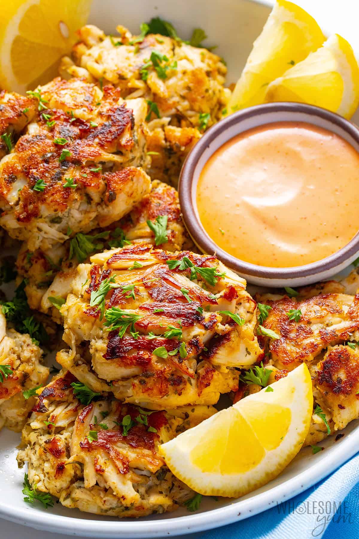 Golden brown Maryland crab cakes with big chunks of crab, on a serving platter with sauce, lemon wedges, and fresh parsley garnish.