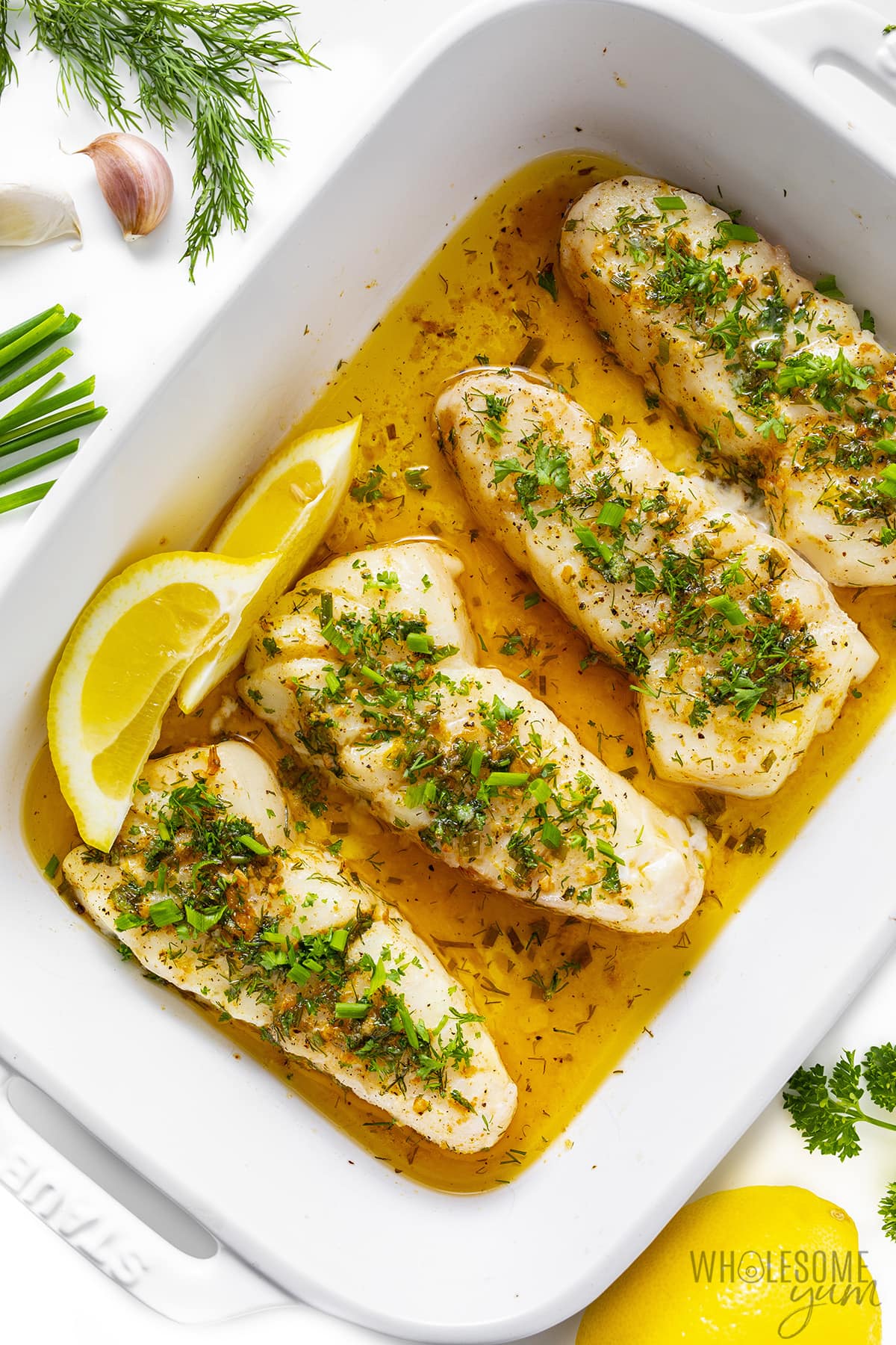 Baked halibut in a baking dish with lemon wedges and fresh herbs.