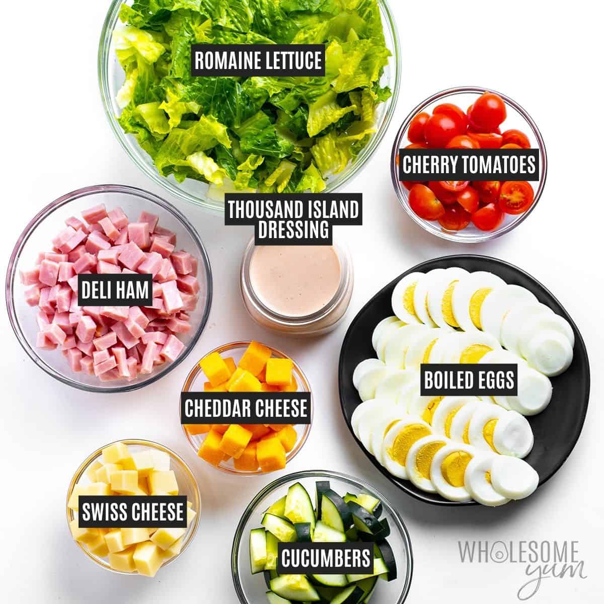 Chef salad ingredients in bowls: Lettuce, ham, cheeses, tomatoes, cucumbers, hard boiled eggs, and thousand island dressing.
