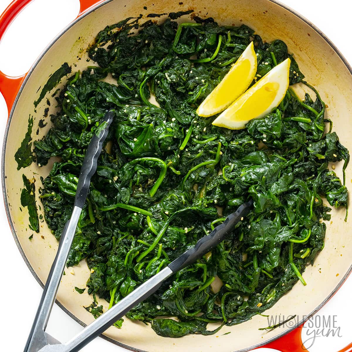 Finished sauteed spinach with garlic, lemon wedges, and tongs.