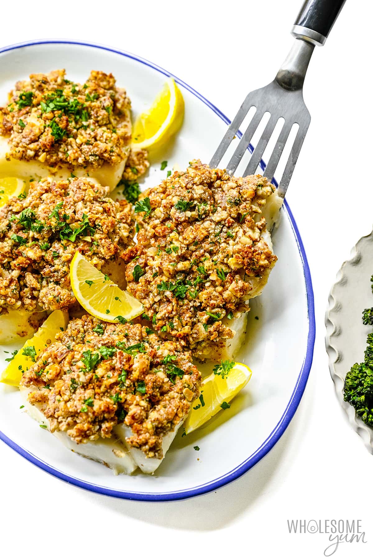 Baked haddock recipe on a plate with almond crust, fish spatula, and lemon wedges.