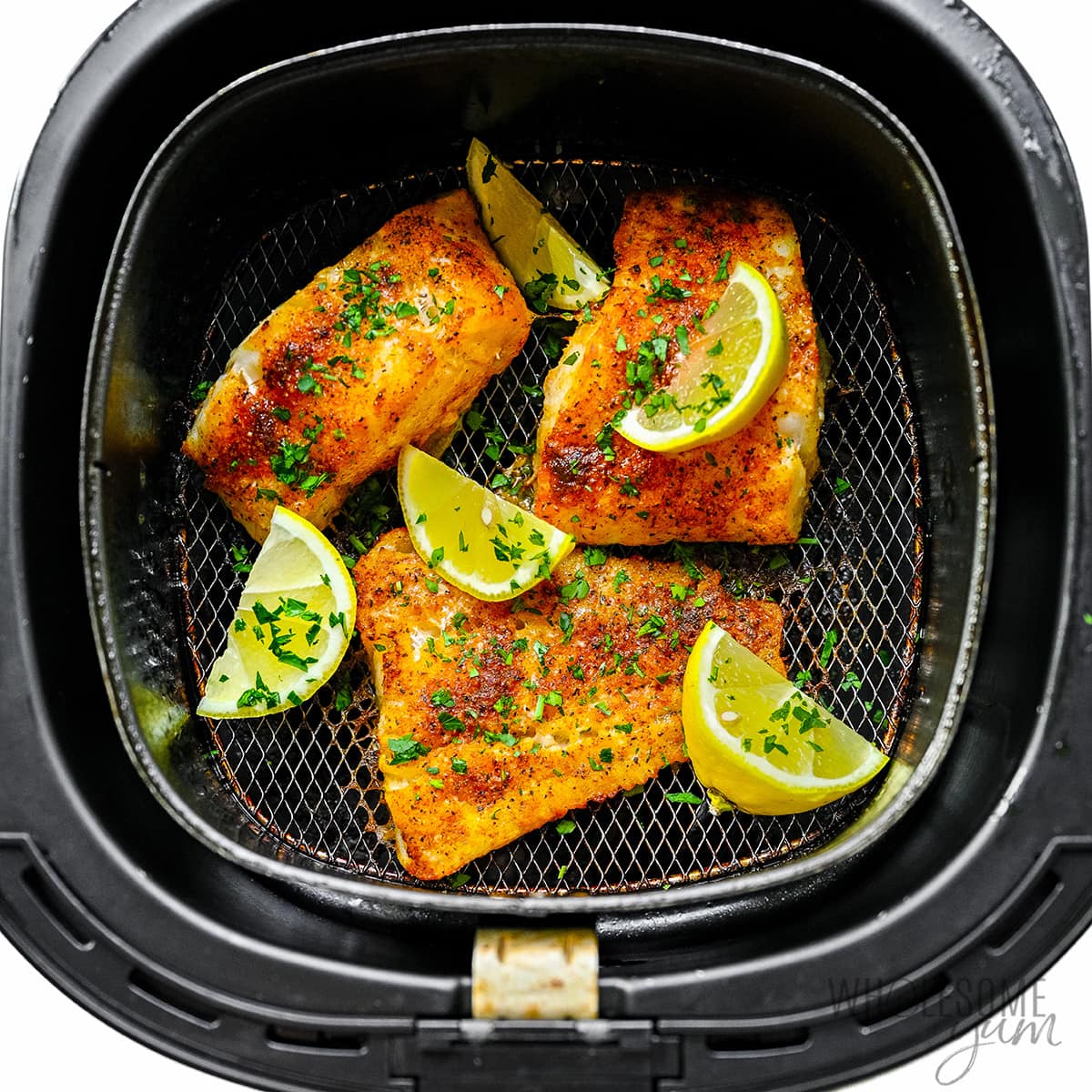 Cooked cod in the air fryer basket with lemon wedges.