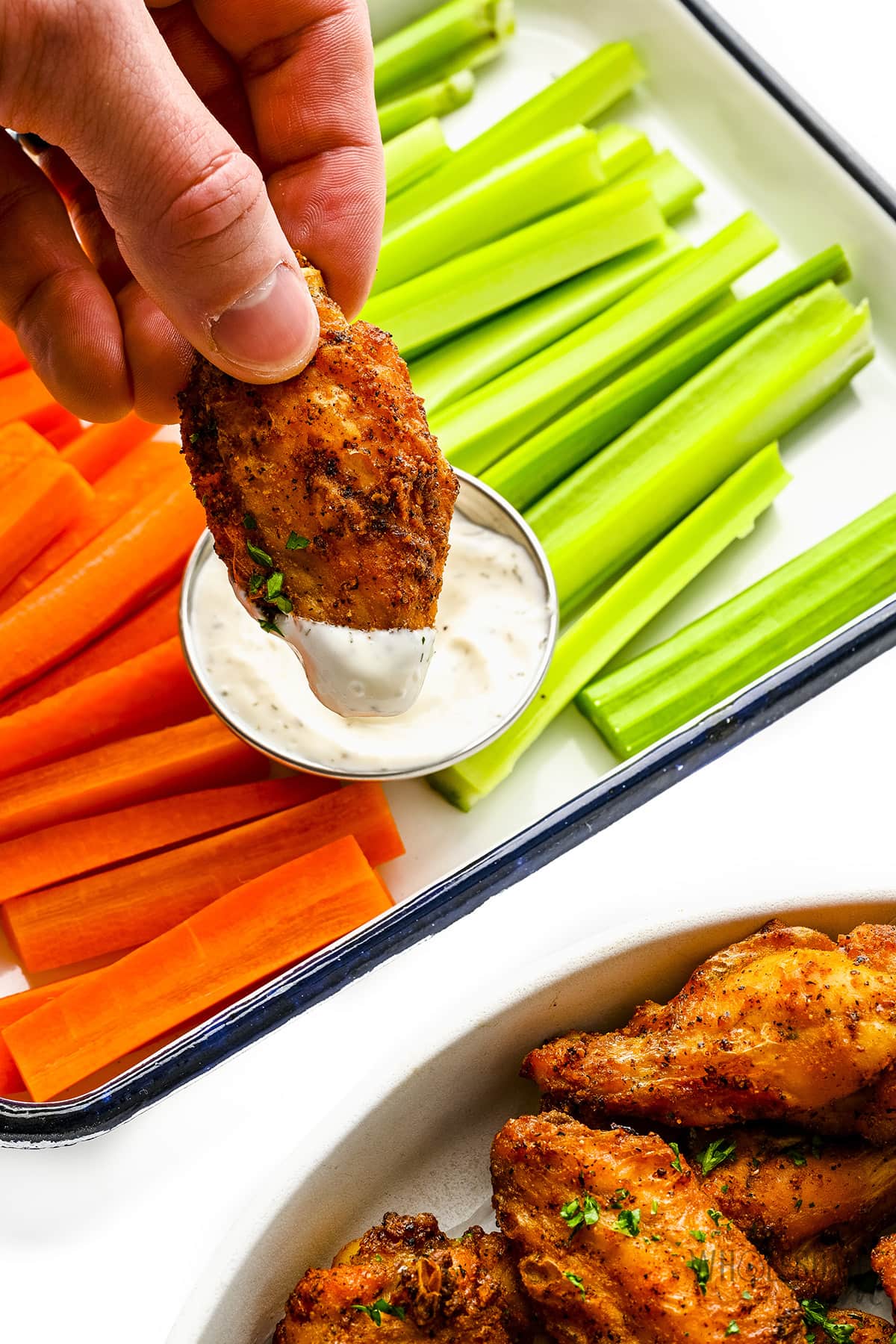 One crispy chicken wing dipped in ranch, with carrots and celery in the background.