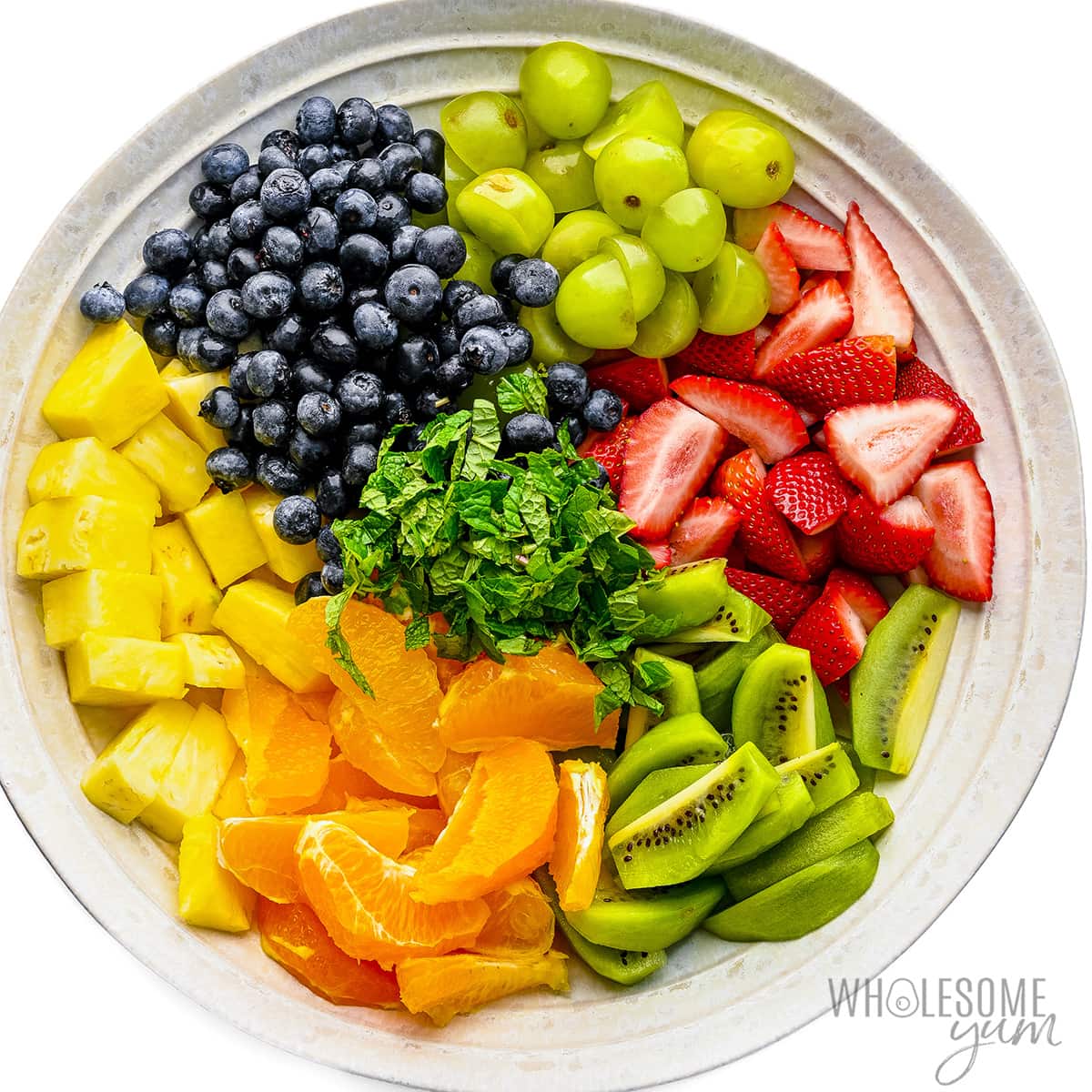 Pineapple, mandarin oranges, kiwi, strawberries, blueberries, grapes, and mint in a bowl.