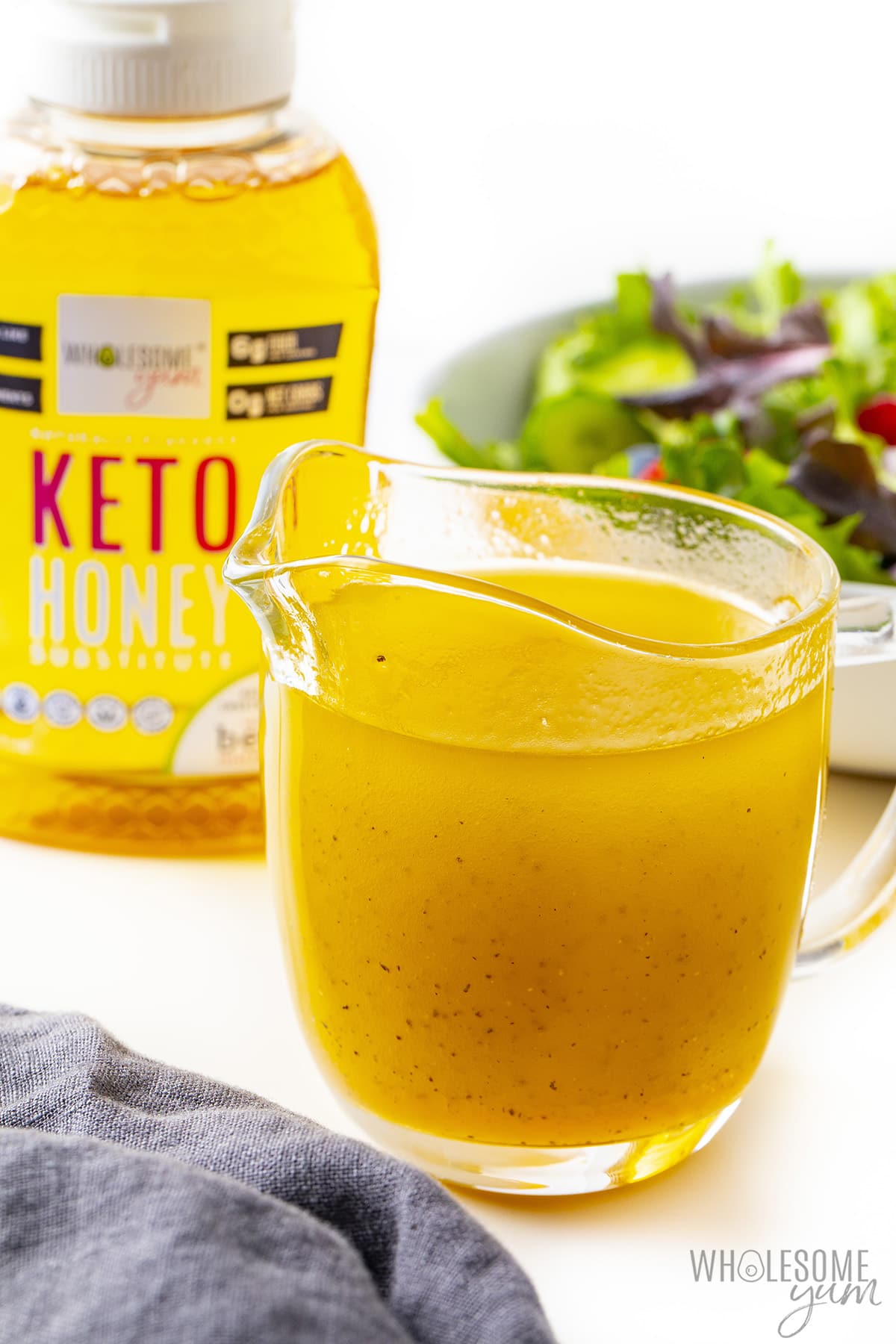 Keto salad dressing in small glass pitcher, with salad and keto honey in the background.