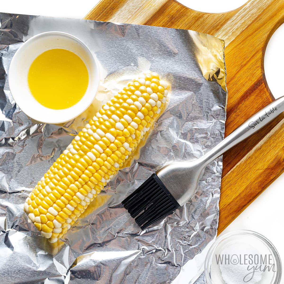 Corn on the cob next to melted butter and brush on aluminum foil.