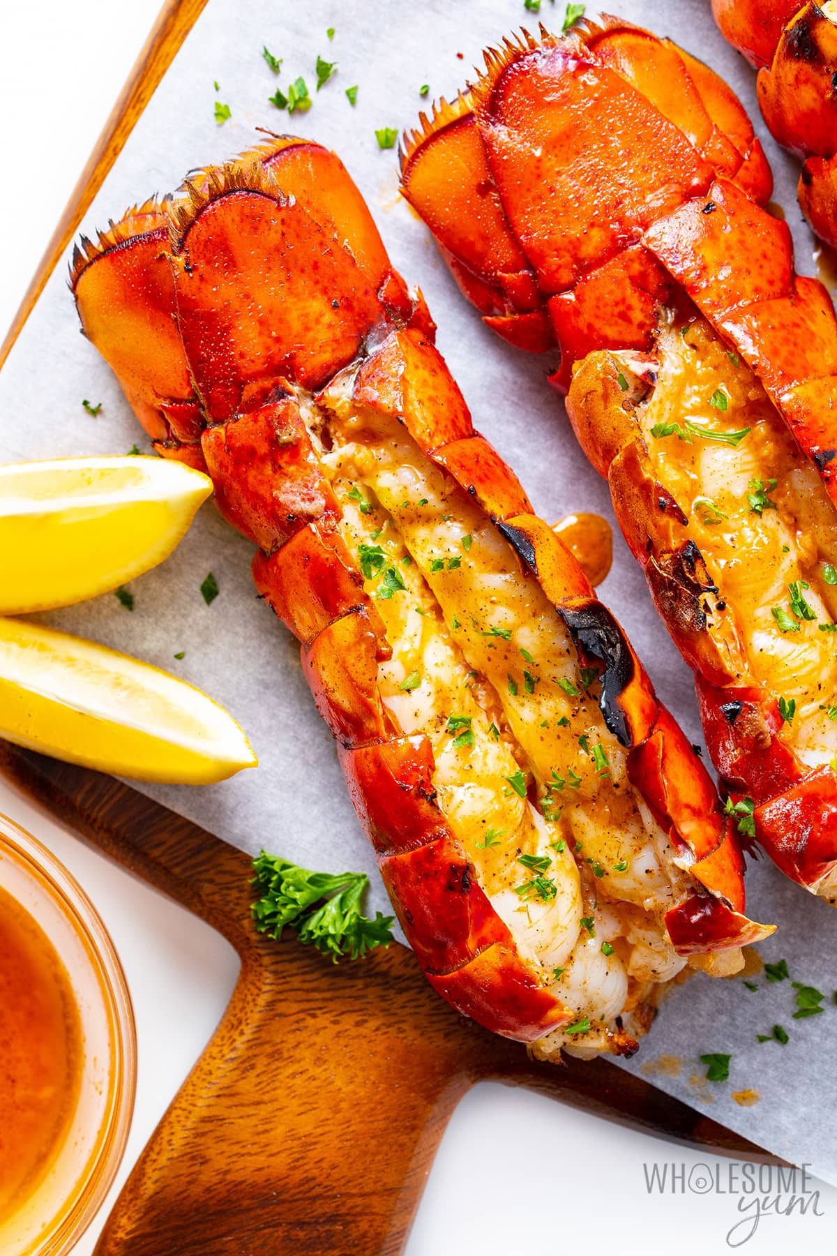 Grilled lobster tails on a cutting board with lemon wedges, fresh parsley, and garlic butter on the side.