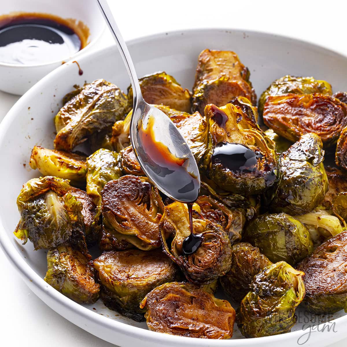 Crispy balsamic brussels sprouts in a bowl, being drizzled with balsamic glaze from a spoon.