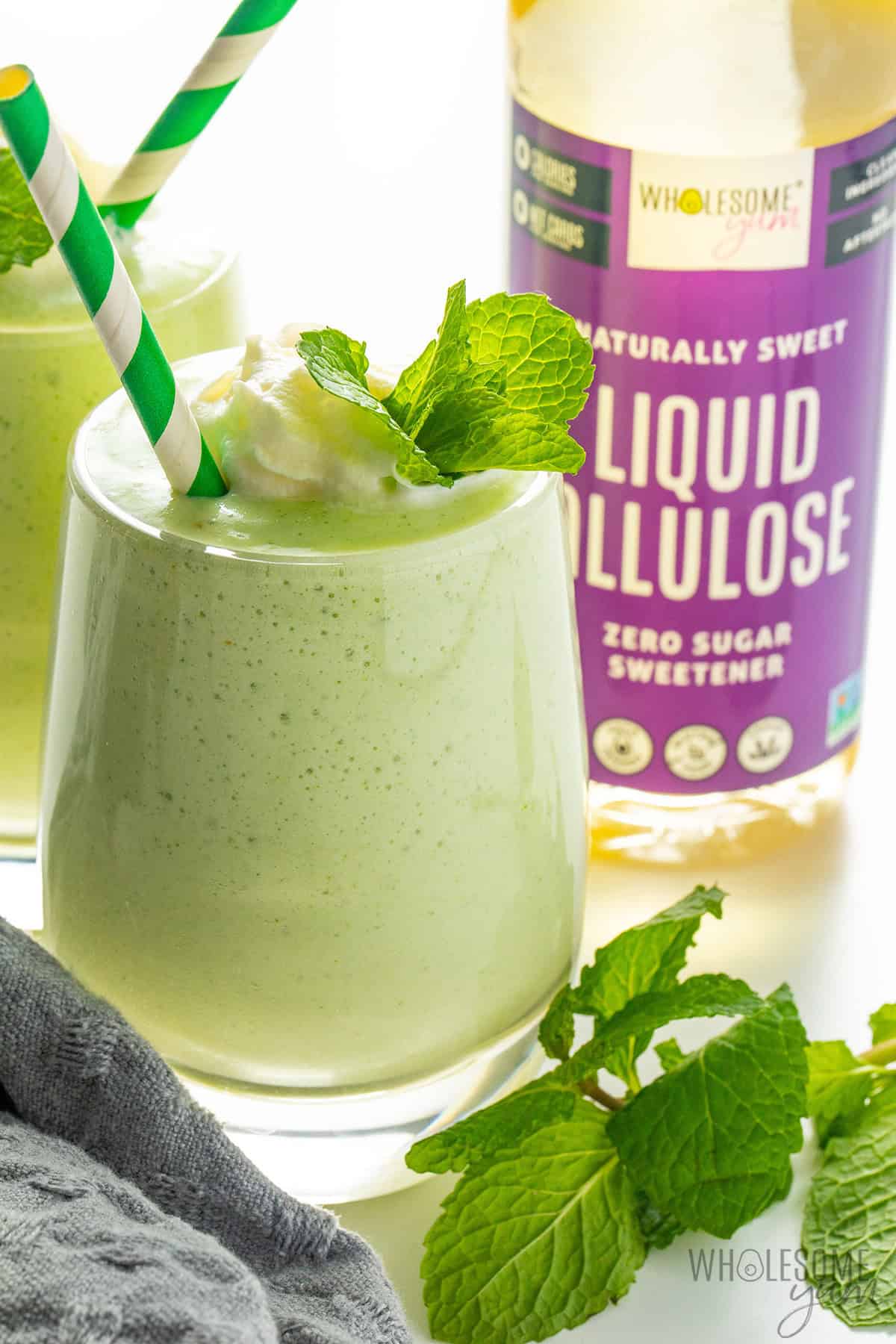 Creamy, healthy shamrock shake in a glass with whipped cream, mint sprig, and a straw, plus liquid allulose in the background.