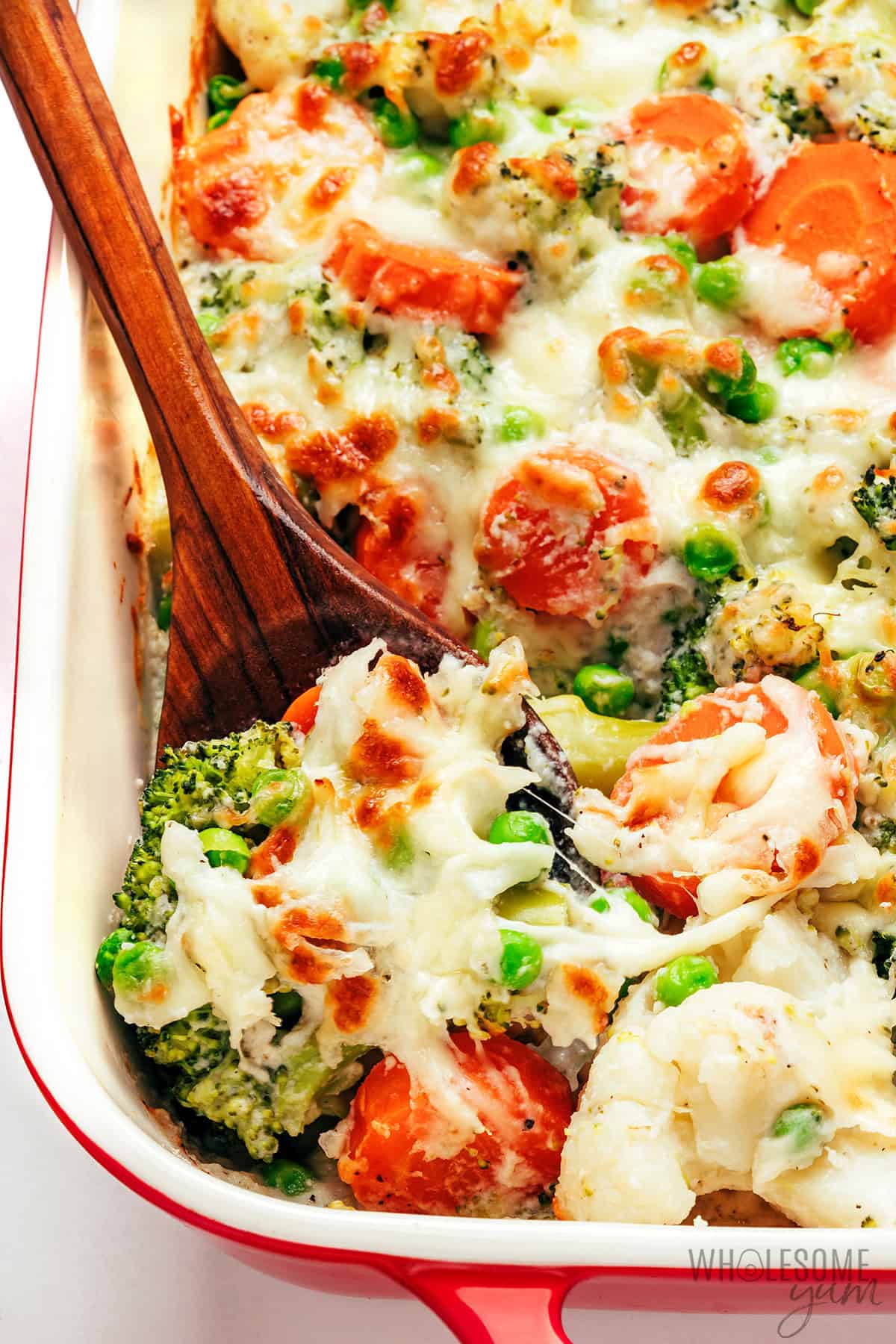 Creamy, cheesy vegetable casserole with broccoli, cauliflower, and carrots, scooped with a wooden spoon.
