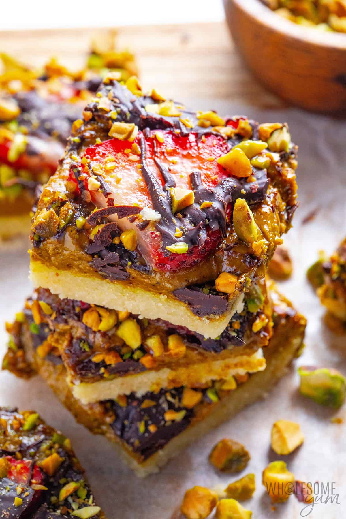 Stack of pistachio chocolate bars with buttery shortbread crust, pistachio cream layer, fresh strawberries, chocolate drizzle, and chopped pistachios.