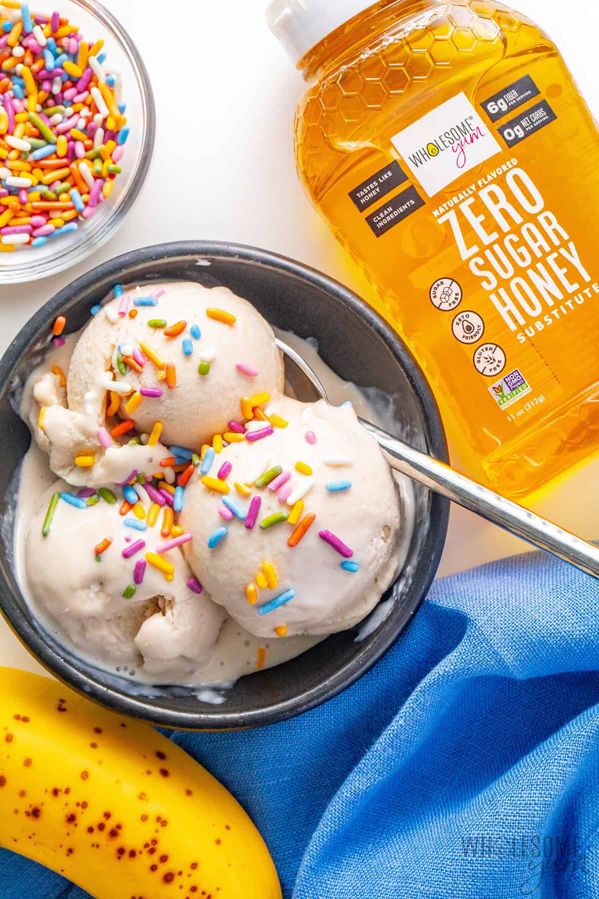 Creamy cottage cheese ice cream in a bowl next to bottle of honey and bowl of sprinkles.