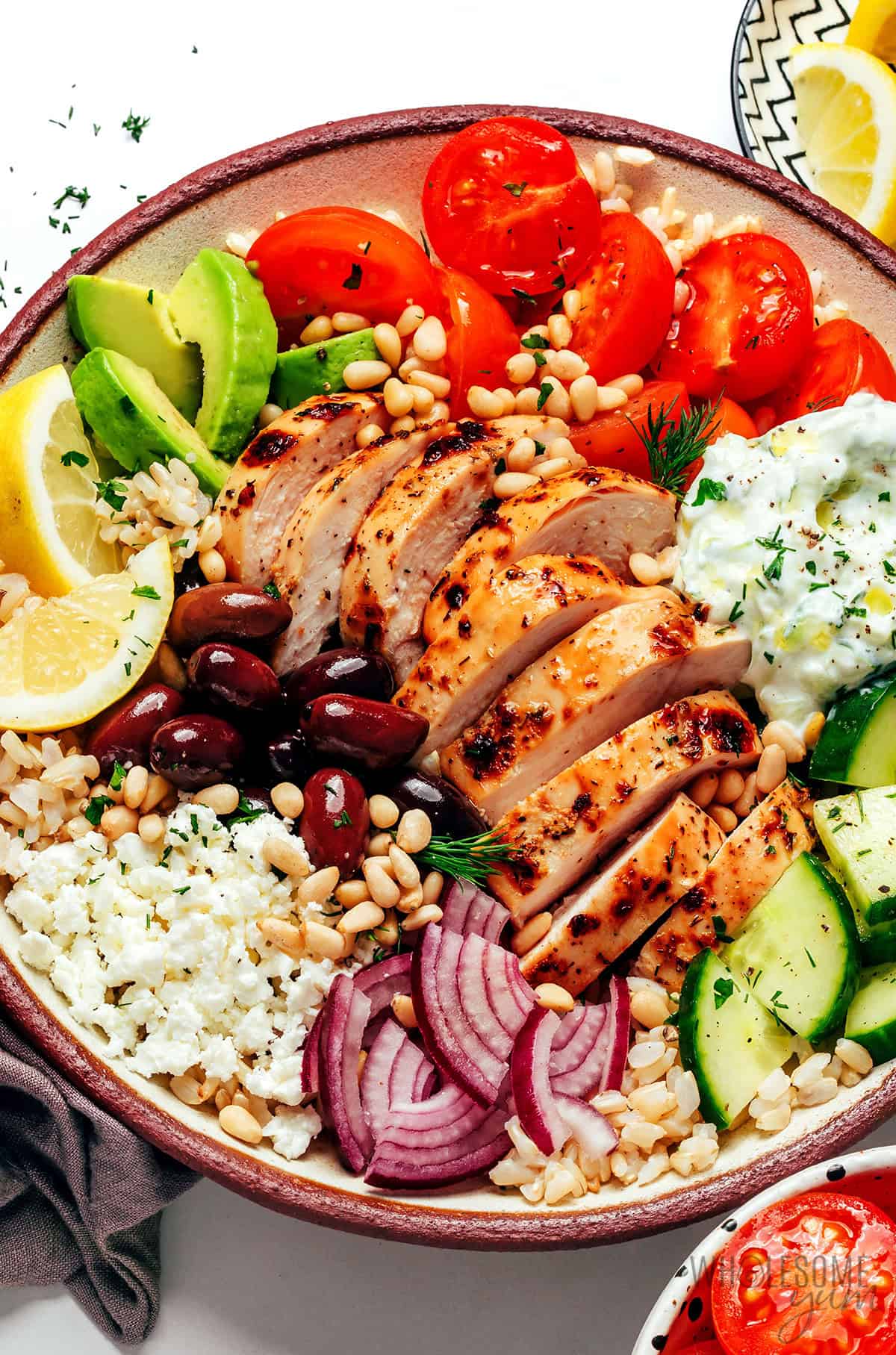 Juicy Greek chicken bowl with veggies, avocado, feta, tzatziki, and pine nuts layered over rice.