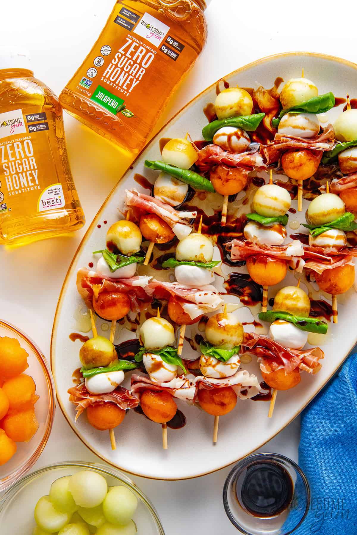 Prosciutto, melon, and mozzarella skewers drizzled with balsamic glaze and honey on a serving plate next to bottles of Wholesome Yum Zero Sugar Honey (original and jalapeno).
