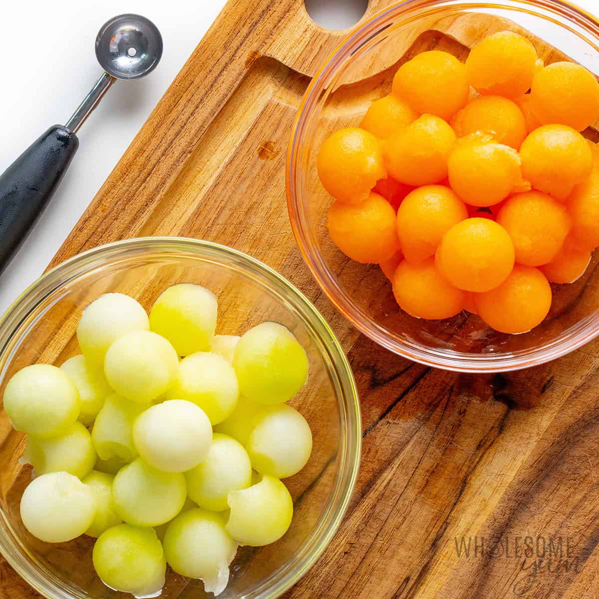 Two bowls filled with cantaloupe and honeydew melon balls on a wooden cutting board.