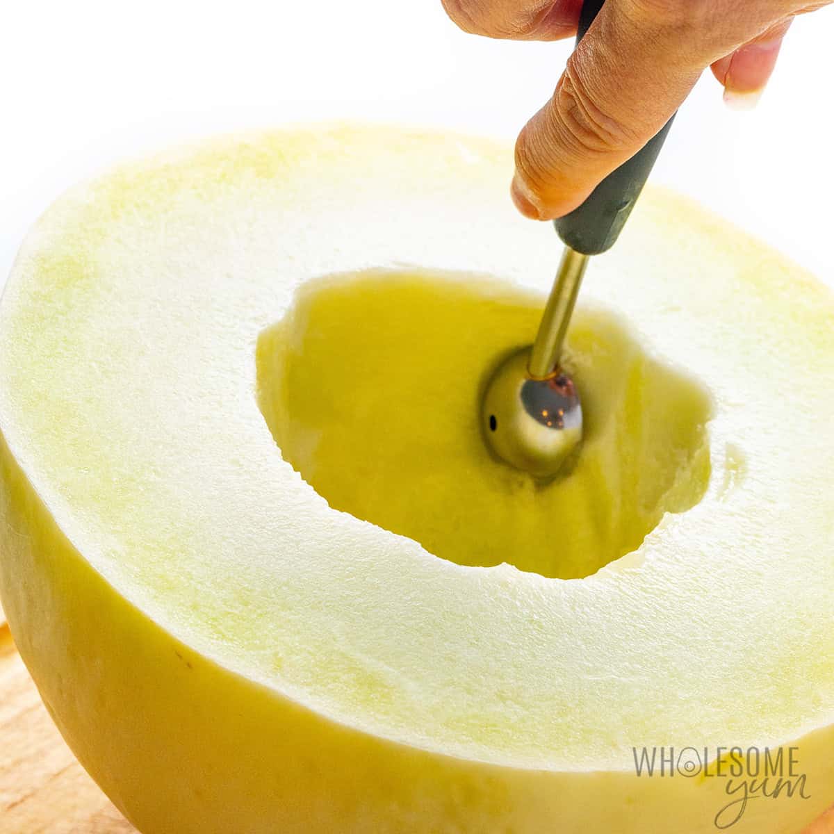 Hand rotating a melon baller in a circular motion inside a honeydew melon.
