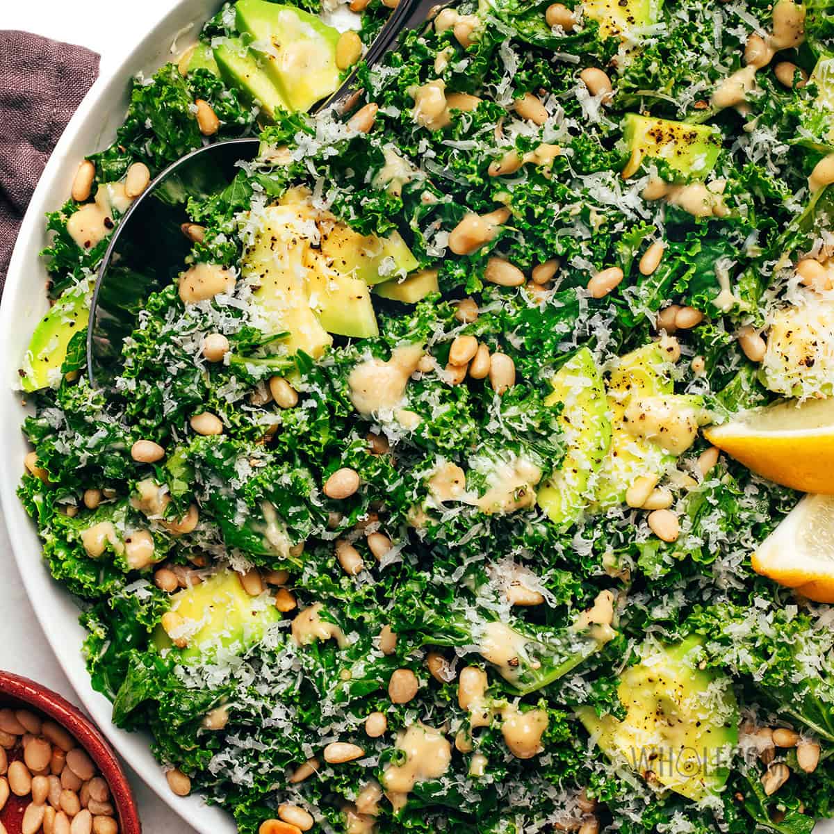 Finished kale Caesar salad recipe with pine nuts, avocado, Parmesan cheese, and lemon wedges.