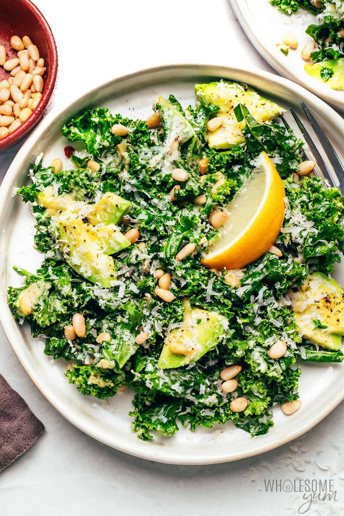 Plated portion of kale Caesar salad topped with a lemon wedge and a bowl of pine nuts.