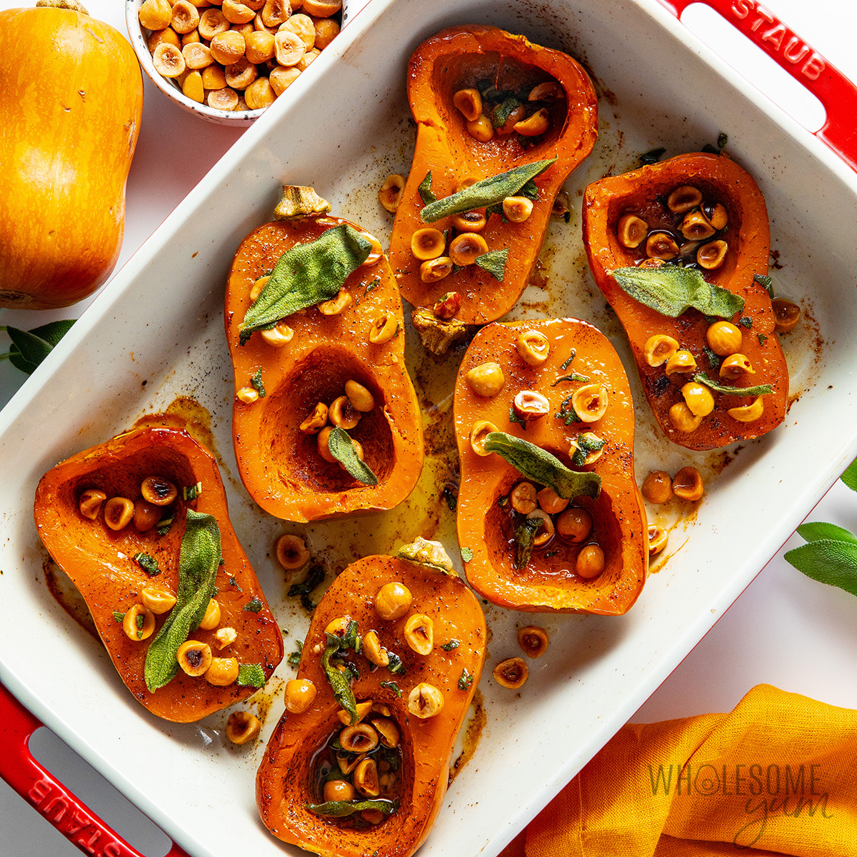 Roasted honeynut squash in a casserole dish topped with hazelnuts and sage leaves.